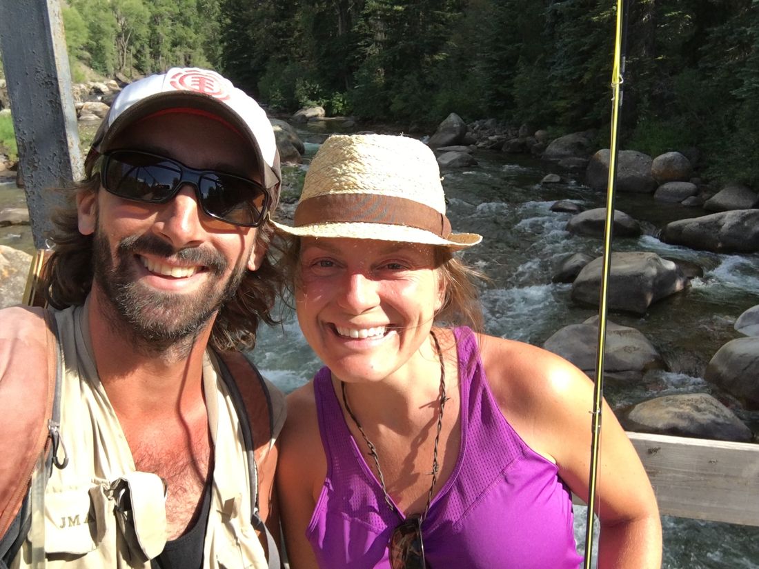 John and Meaghan smiling on a footbridge over a rocky mountain creek, fly rod in hand